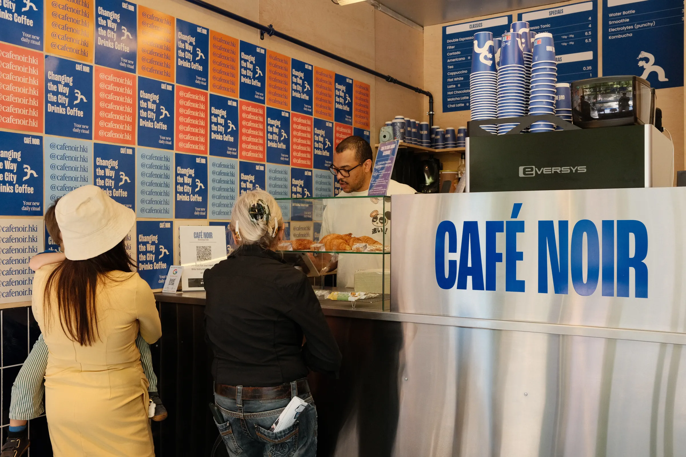 Customers ordering at the Café Noir counter under walls of blue and orange posters